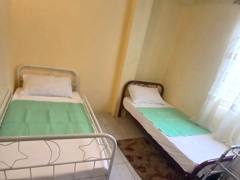 A bright shared bedroom with two single beds made up with white linens, a rug between them and a walking frame beside one bed