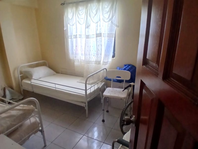 A shared bedroom seen from the doorway: a single bed with safety rails beneath a curtained window, with a shower chair and a walking frame nearby